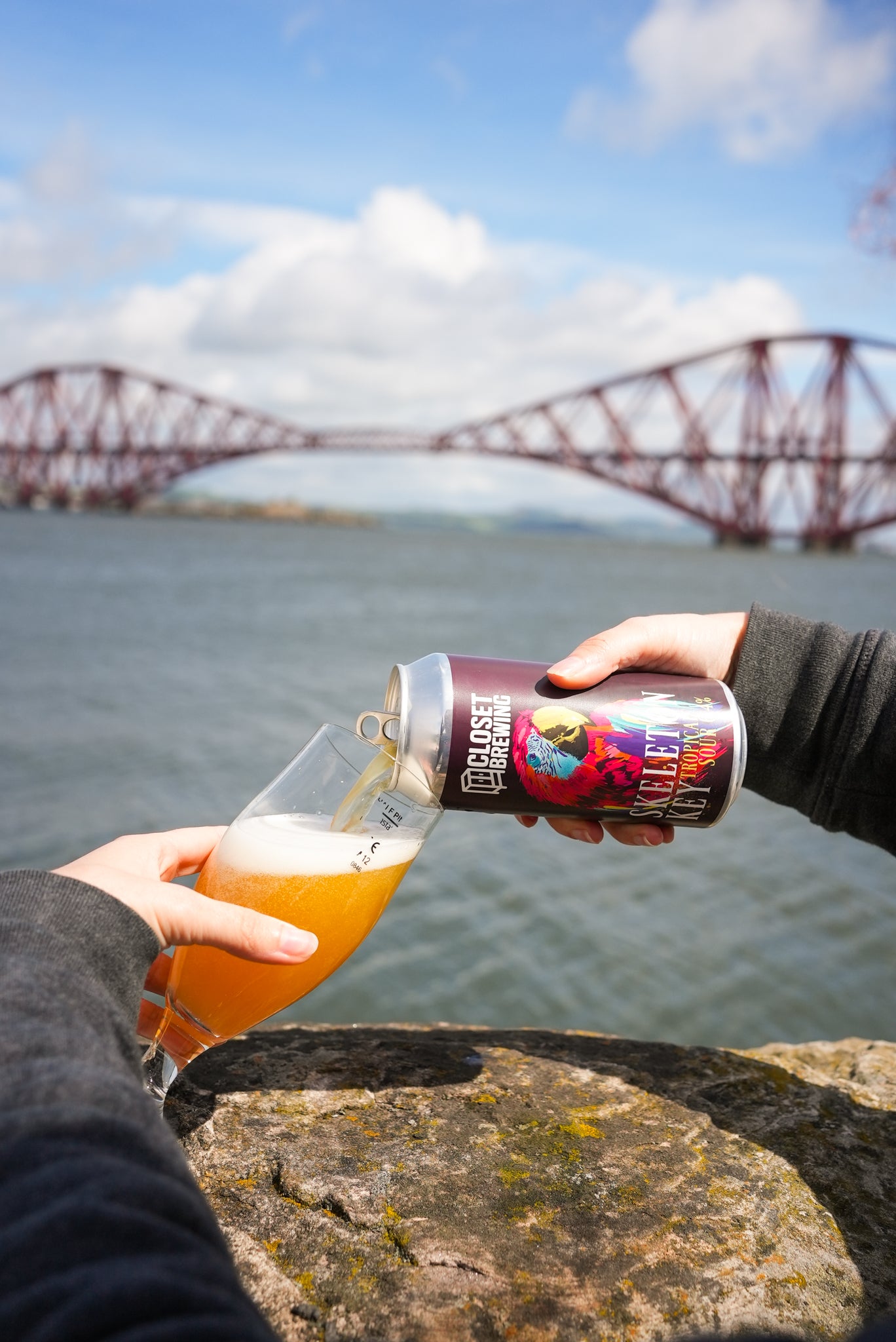 Person pouring beer from a can into a glass with a bridge and water in the background